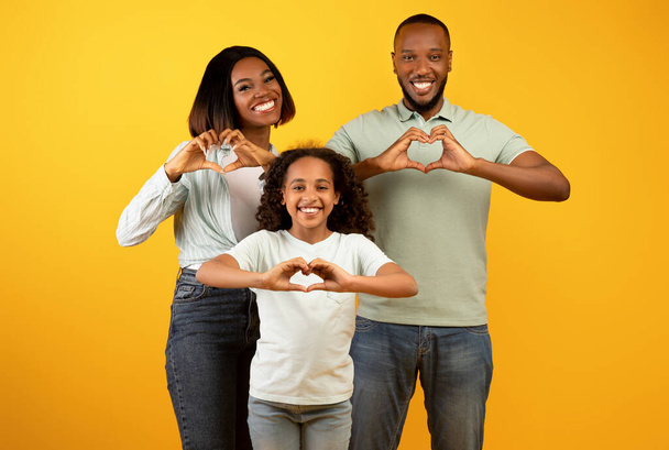 Kochająca rodzina. Szczęśliwy Afryki amerykańskich rodziców i liitle córka pokazując znaki kształt serca na żółtym tle - Zdjęcie, obraz