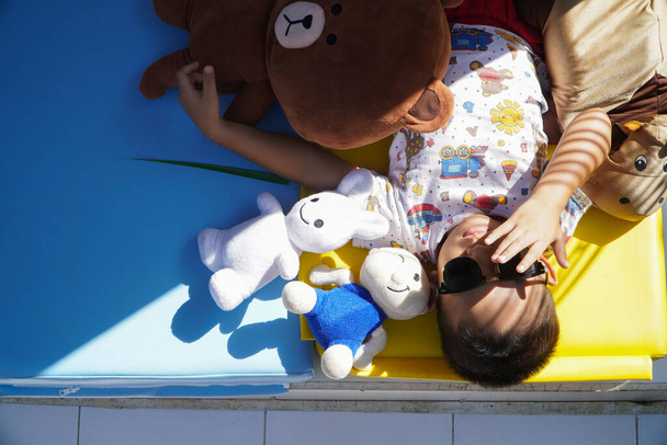 Een peuter ligt te zonnebaden met zijn favoriete poppen op een gekleurde matras. Hij lijkt te genieten van de zon in een poging om een natuurlijke bron van vitamine D te krijgen van zonlicht.. - Foto, afbeelding