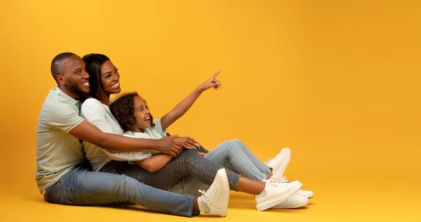 Alegre familia afroamericana sentada en el suelo, linda chica apuntando con el dedo al espacio de copia sobre fondo amarillo, panorama - Foto, Imagen
