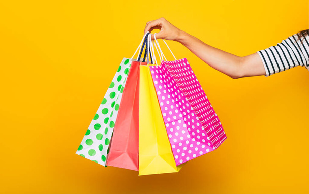 Close up photo of female hand with colorful shopping bag in hands isolated on yellow background - Zdjęcie, obraz