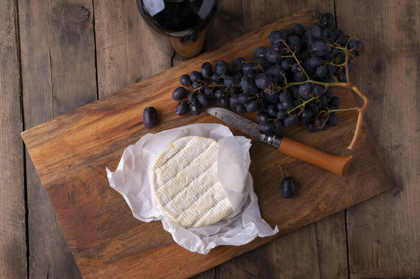 A round piece of cheese wrapped in a white napkin on a wooden board and grapes with a glass of wine. - Valokuva, kuva