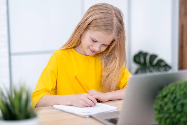 Blonde schoolgirl studying at home doing school homework with laptop. Distance learning online education. Back to school - Zdjęcie, obraz