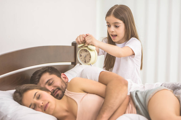 A lány egy ébresztőórát tart a szülők közelében az ágyban. - Fotó, kép