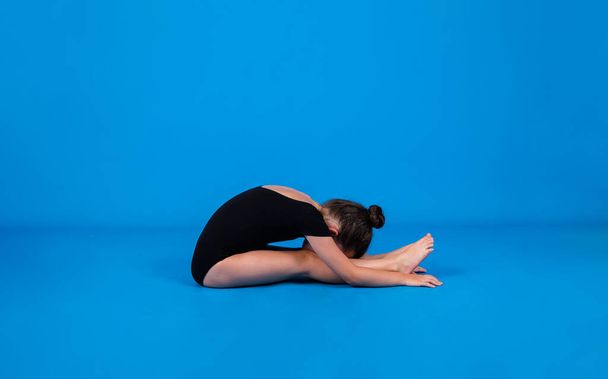 Маленькая гимнастка в черном купальнике выполняет упражнения пластичности на синем фоне с местом для текста - Фото, изображение