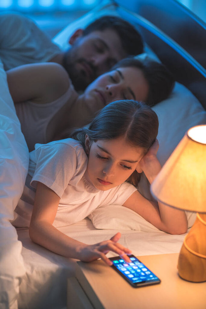 Телефон маленькой девочки рядом с родителями в кровати. ночь время - Фото, изображение