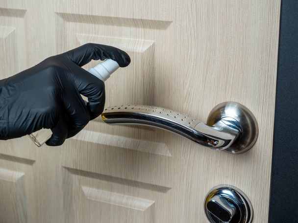 Человек в черных латексных перчатках брызгает антисептиком на дверную ручку. Обработка элементов, чтобы уменьшить вероятность заражения вирусами - Фото, изображение