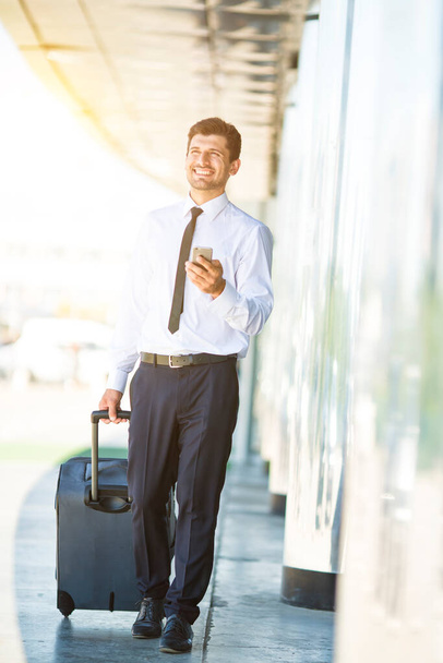 De gelukkige zakenman telefoon en lopen met een koffer in de buurt van gebouw - Foto, afbeelding