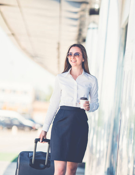 Деловая женщина в солнечных очках держит чашку кофе и ходит с чемоданом - Фото, изображение