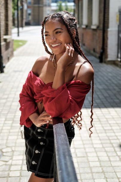 Молодая женщина с классными волосами позирует на лондонской улице в жаркий летний день. У нее косички и красный топ.. - Фото, изображение
