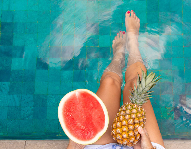 Summer concept. Female legs in the pool, in the hands of pineapple and watermelon - Zdjęcie, obraz