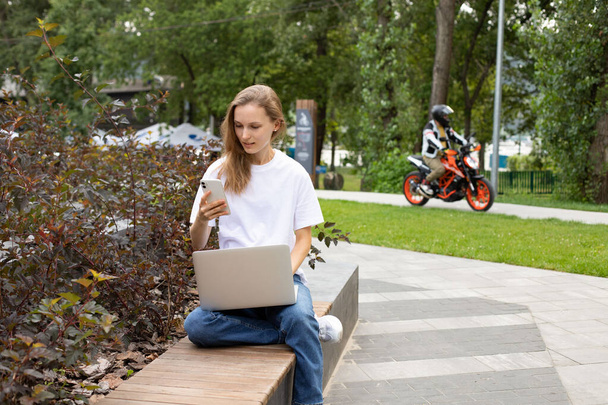 Νεαρή φοιτήτρια Caucaisan στέλνοντας μηνύματα sms κινητής τηλεφωνίας κατά τη διάρκεια του χρόνου για e-learning στο πάρκο στον πάγκο, χιλιετή hipster κορίτσι σε μοντέρνα φθορά σύνδεση με bluetooth για την ανταλλαγή αρχείων πολυμέσων σε ψηφιακό netbook - Φωτογραφία, εικόνα