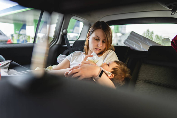 Ydult femme caucasienne mère assise sur le siège arrière de sa voiture tenant formule ou lait maternel dans le biberon prêt à nourrir son enfant tout en voyageant sur la route en été jour en vacances - Photo, image