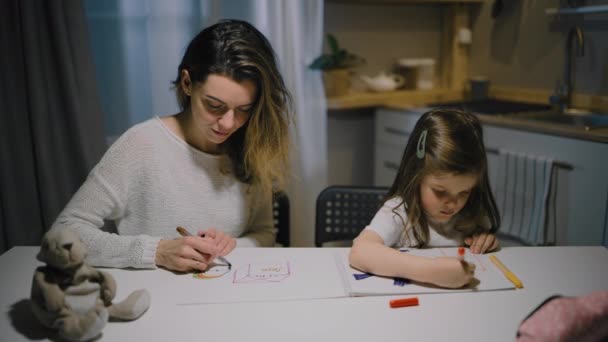 Una madre y una hija pequeña dibujan dibujos en papel en la cocina con marcadores - Imágenes, Vídeo