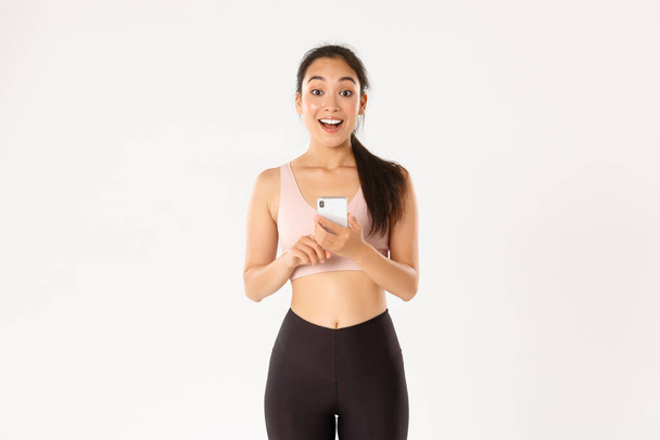 Spor, teknoloji ve aktif yaşam tarzı konsepti. Şaşırmış ve şaşırmış Asyalı bayan atlet, sporcu kadın diyor ki vay canına, cep telefonundaki fitness uygulamasını kontrol etmek, egzersiz takip cihazı kullanmak, uygulamayla çalışmak - Fotoğraf, Görsel