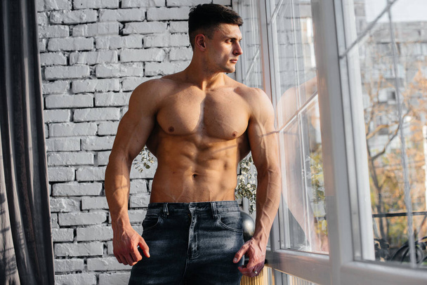 Młody seksowny sportowiec z idealnym brzuchem pozuje przy oknie w studio topless w dżinsach na tle. Zdrowy styl życia, prawidłowe odżywianie, programy treningowe i odżywianie dla utraty wagi. - Zdjęcie, obraz