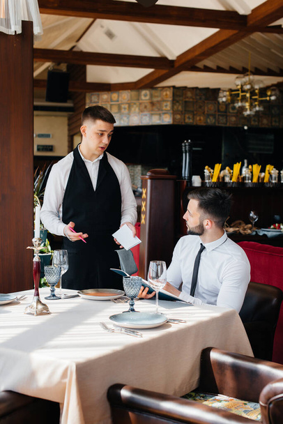 A young businessman in a fine restaurant examines the menu and makes an order to a young waiter in a stylish apron. Customer service. - Foto, imagen