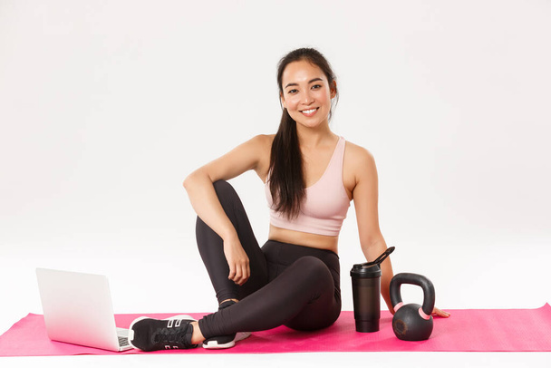Portrait of smiling cute asian fitness girl staying at home during covid-19 social distancing, sitting on rubber mat for workout, use laptop for online training with coach, repeat after video - Photo, Image