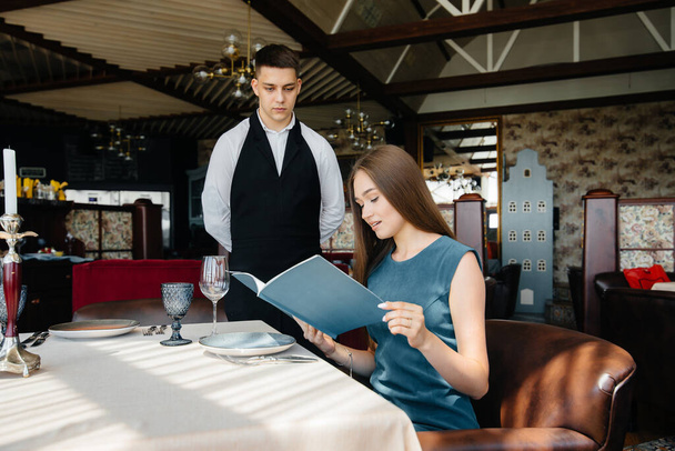 A young beautiful woman in a fine restaurant looks at the menu and makes an order to a young waiter in a stylish apron. Customer service. - Photo, Image