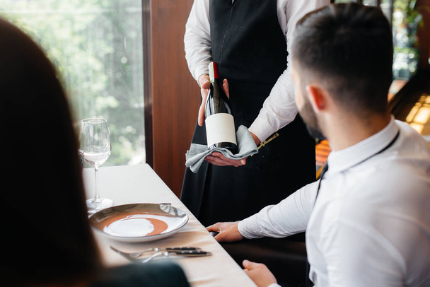 A young waiter in a stylish apron demonstrates and offers a fine wine to a beautiful couple in a restaurant. Customer service. - Φωτογραφία, εικόνα