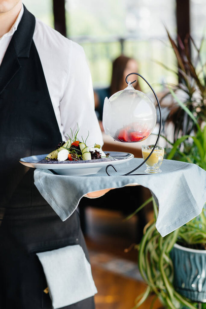 A young waiter in a stylish uniform stands with an exquisite dish on a tray near the table in a beautiful restaurant close-up. Restaurant activity, of the highest level. - 写真・画像