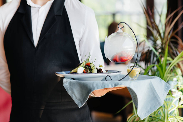 A young waiter in a stylish uniform stands with an exquisite dish on a tray near the table in a beautiful restaurant close-up. Restaurant activity, of the highest level. - Foto, imagen
