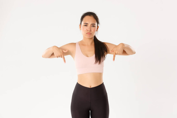 Sport, wellbeing and active lifestyle concept. Upset and disappointed asian sportswoman, woman in fitness clothing pointing fingers down with sulking face, complaining on banner, white background - Photo, Image