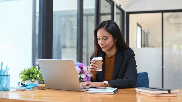 Щаслива успішна бізнес-леді п'є гарячу каву вранці і читає новини на ноутбуці комп'ютера
. - Фото, зображення
