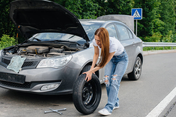 A young girl stands near a broken car in the middle of the highway and tries to change a broken wheel on a hot sunny day. Failure and breakdown of the car. Waiting for help. - Фото, зображення