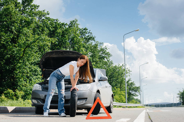 A young girl stands near a broken car in the middle of the highway and tries to change a broken wheel on a hot sunny day. Failure and breakdown of the car. Waiting for help. - Foto, afbeelding
