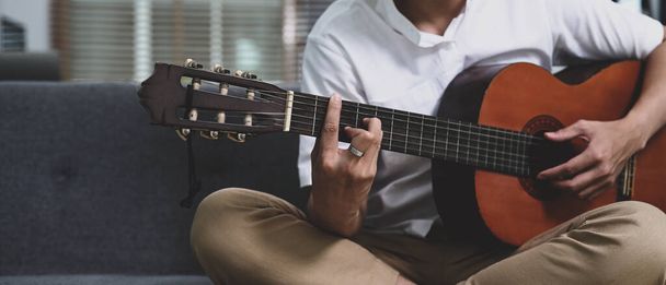 Cropped shot casual man playing guitar while sitting on sofa in living room. - Foto, immagini