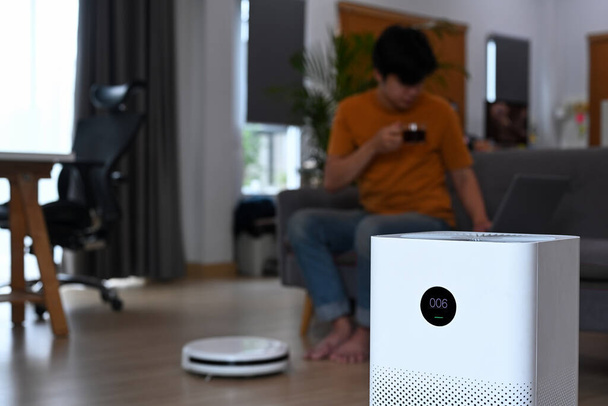 Air purifier on wooden floor in living room and man drinking coffee on sofa. - Photo, Image