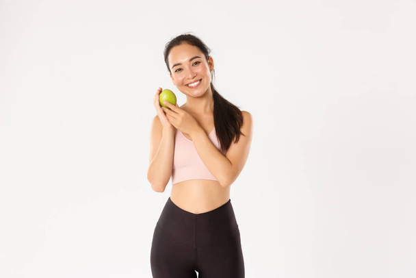 Sport, wellbeing and active lifestyle concept. Smiling cute asian fitness coach, girl athlete in sportswear showing green apple, sit on diet and workout to gain perfect body, white background - Photo, Image