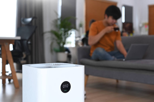 Air purifier on the floor in living room and young man sitting on sofa in background. - Photo, Image
