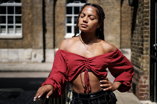 Young woman with cool hair posing in a street of London in a hot summer day. She wears braids and red top. - Photo, Image