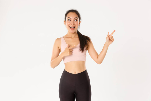 Sport, wellbeing and active lifestyle concept. Excited slim and cute asian fitness girl, female athlete pointing fingers upper right corner, showing workout equipment sale promo or gym offer - Foto, Imagem