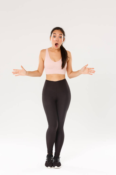 Full length of astonished and surprised female athlete, sportswoman in activewear looking impressed, drop jaw and spread hands sideways see great gym discount, workout equipment in sale - Foto, Imagem