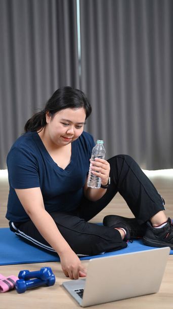 Happy obese woman holding water bottle and watching fitness training video on laptop at home. - Fotografie, Obrázek