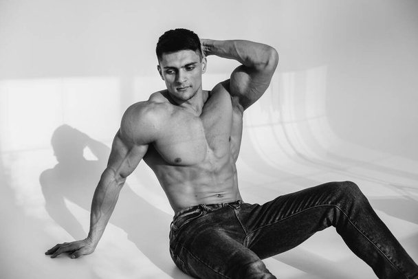 A young sexy athlete with perfect abs is sitting in the studio topless in jeans. Healthy lifestyle, proper nutrition, training programs and nutrition for weight loss. Black and white. - Foto, Imagem