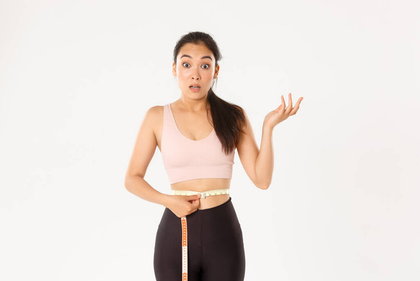 Fitness, healthy lifestyle and wellbeing concept. Surprised asian girl on diet, sportswoman wrap measuring tape around waist and look impressed as lose weight with workout - Photo, Image