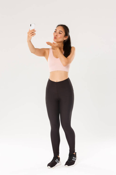 Πλήρες μήκος του χαριτωμένο και ανόητο γυναίκα blogger στο γυμναστήριο, φορώντας αθλητικά ρούχα και τη λήψη selfie ή ζωντανή ροή από γυμναστήριο προπόνηση συνεδρία, στέλνοντας φιλί αέρα στην οθόνη του κινητού τηλεφώνου, στέκεται λευκό φόντο - Φωτογραφία, εικόνα