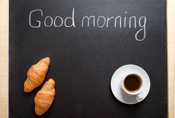 Una taza de café, croissants, la inscripción "Buenos días" en una pizarra negra. Un desayuno tradicional, buen comienzo del día concepto. Copiar espacio para el menú. - Foto, imagen