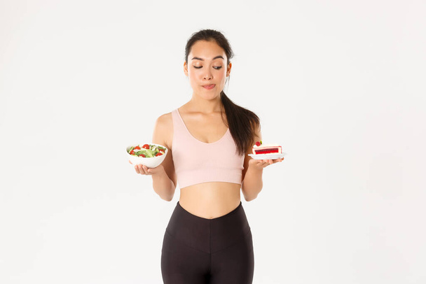 Active lifestyle, fitness and wellbeing concept. Portrait of tempting cute asian girl trying resist temptation as looking at delicious cake, being on diet, need eat healthy salad, licking lips - Photo, Image