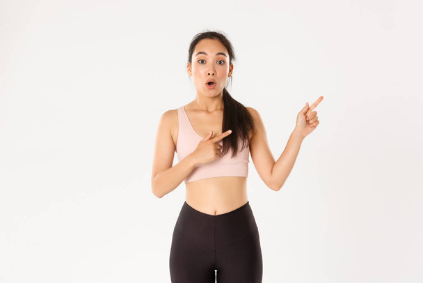 Sport, wellbeing and active lifestyle concept. Surprised and amazed asian girl in fitness clothing, pointing fingers upper right corner, gasping and say wow impressed with gym discounts offer - Photo, image