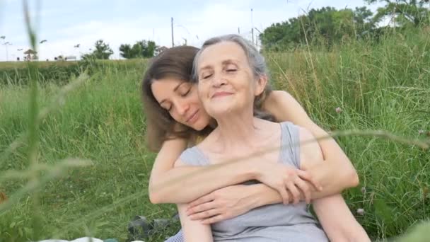 Vanhempi äiti harmailla hiuksilla aikuisen tyttärensä kanssa puutarhassa halaamassa toisiaan piknikillä aurinkoisena päivänä ulkona, äitienpäivänä - Materiaali, video