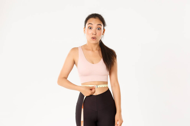 Fitness, zdravý životní styl a koncept pohody. Překvapená asijské dívka na dietě, sportovkyně zábal měřicí pásku kolem pasu a vypadají ohromeně, jak zhubnout s cvičením - Fotografie, Obrázek