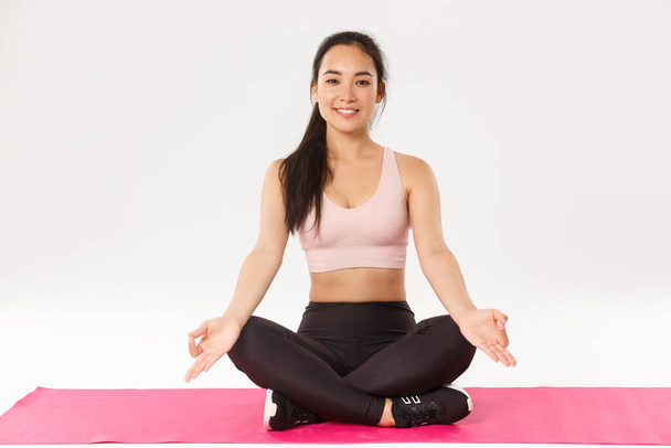 Portret uśmiechniętej, zrelaksowanej i spokojnej azjatyckiej brunetki z idealnym ciałem, wyglądającej szczęśliwie, siedzącej w pozycji lotosu, skrzyżowanych nóg na gumowej macie do ćwiczeń fitness lub jogi, medytującej, białe tło - Zdjęcie, obraz