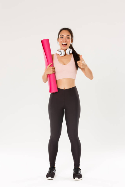 Full length of cheerful satisfied, slim asian girl with fitness rubber mat and headphones, female athlete showing thumbs-up, recommend gym or yoga classes, looking pleased after good workout - Фото, зображення