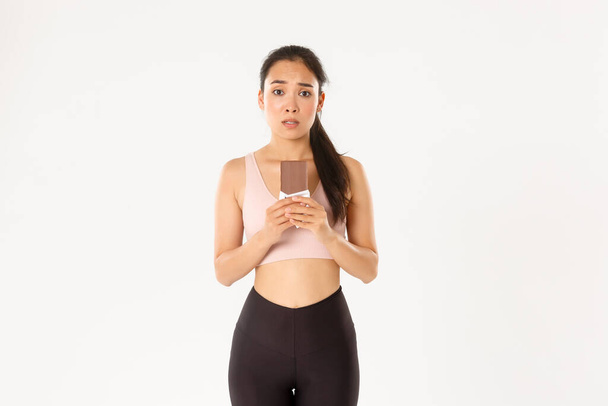 Sport, wellbeing and active lifestyle concept. Indecisive cute asian girl in workout clothing, looking hesitant at chocolate bar, trying resist temptation as losing weight on diet - Photo, Image