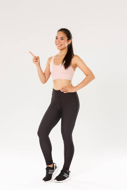 Sport, gym and healthy body concept. Full length smiling satisfied female athlete, asian fitness girl in active wear making her choice, pointing and looking upper left corner, pick gym for training - Fotografie, Obrázek