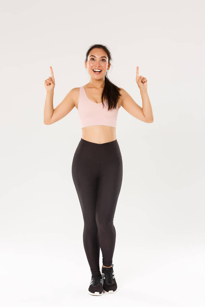 Sport, gym and healthy body concept. Full length of excited smiling female athlete in sportswear looking amazed and pointing fingers up, female athlete enjoying fitness, invite new members - Foto, Imagem